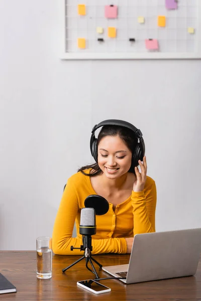Joven asiático radio host tocando auriculares inalámbricos mientras está sentado cerca de micrófono, portátil y teléfono inteligente con pantalla en blanco - foto de stock