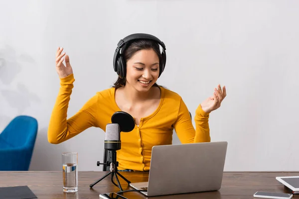 Excited asian announcer in wireless headphones showing wow gesture near microphone and laptop — Stock Photo