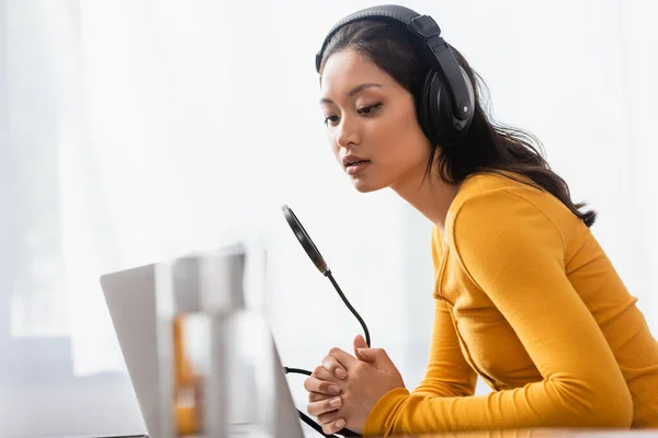 Enfoque selectivo de reflexivo asiático locutor en auriculares inalámbricos sentado con las manos cerradas cerca del micrófono - foto de stock