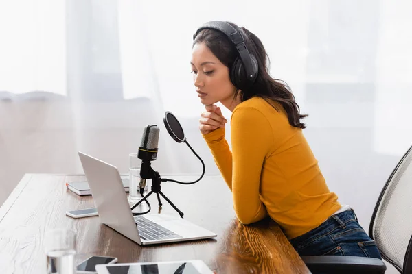 Thoughtful asian radio host in wireless headphones touching chin while sitting at workplace in studio — Stock Photo