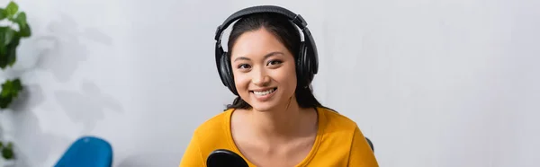 Горизонтальное изображение радостного азиатского вещателя в беспроводных наушниках, смотрящего на камеру в студии радио — стоковое фото
