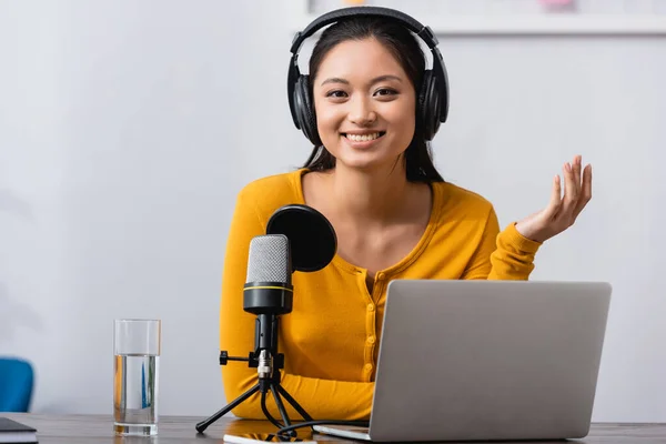 Aufgeregter asiatischer Radiomoderator mit drahtlosen Kopfhörern blickt in die Kamera, während er neben Mikrofon und Laptop sitzt — Stockfoto