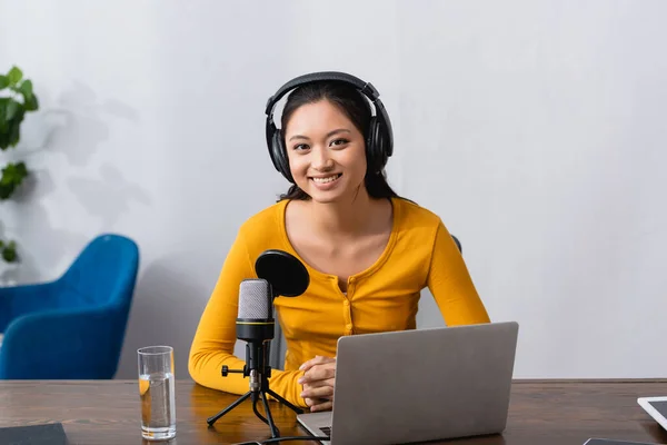 Freudiger asiatischer Ansager mit drahtlosen Kopfhörern, der in die Kamera blickt, während er neben Laptop und Mikrofon sitzt — Stockfoto