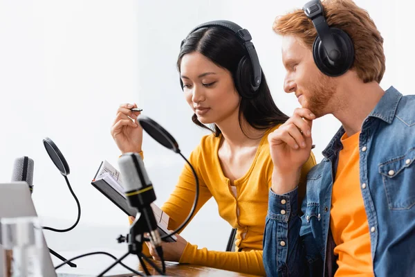Interracial couple of broadcasters in wireless headphones looking at notebook near microphones in studio — Stock Photo