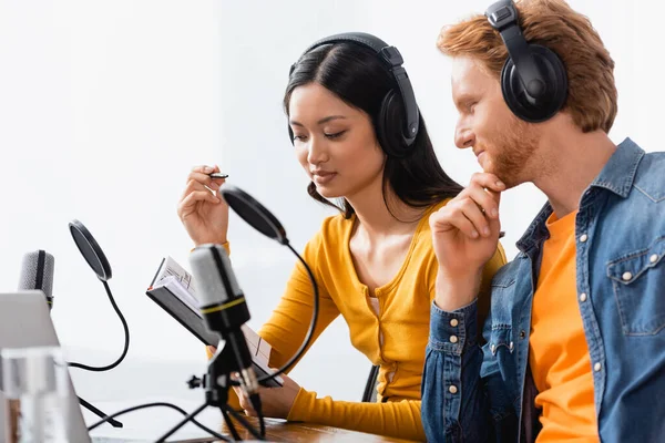 Pareja interracial de emisoras en auriculares inalámbricos mirando portátil cerca de micrófonos en el estudio - foto de stock