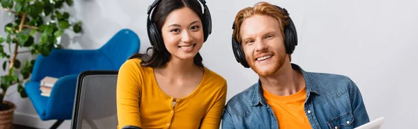 Tiro panorâmico de casal interracial de anunciantes alegres em fones de ouvido sem fio olhando para a câmera no estúdio de rádio — Fotografia de Stock