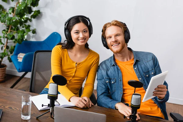 Joyeuses animateurs de radio multiculturelles dans les écouteurs sans fil regardant la caméra tandis que l'homme tenant tablette numérique — Photo de stock