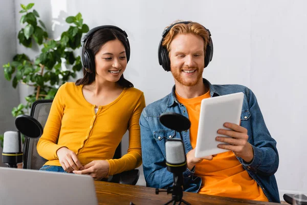 Radiodiffuseur interracial excité dans les écouteurs sans fil regardant tablette numérique sur le lieu de travail — Photo de stock