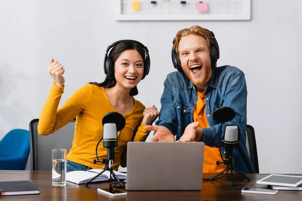 Excited broadcaster pointing with hands at laptop near asian colleague showing triumph gesture — Stock Photo