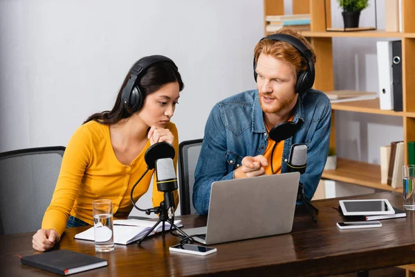 Hôte radio pointant avec le doigt à l'ordinateur portable près collègue asiatique réfléchi dans les écouteurs sans fil — Photo de stock