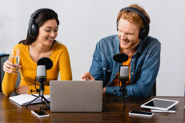 Young announcer pointing with finger at laptop near asian colleague holding glass of water — Stock Photo