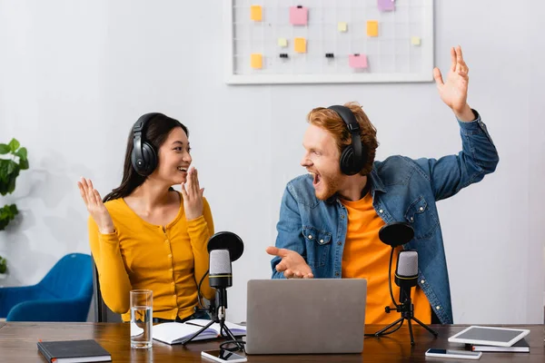 Excited multicultural broadcasters in wireless headphones gesturing and looking at each other at workplace — Stock Photo