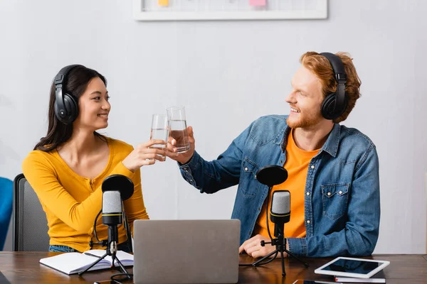 Interracial couple of young radio hosts clinking glasses of water at workplace — Stock Photo