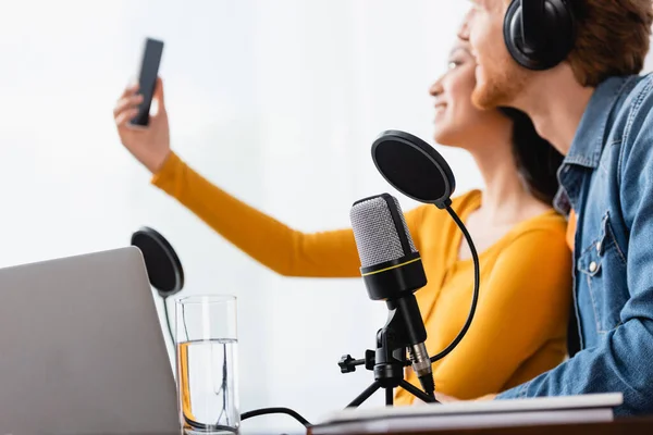 Selective focus of asian announcer taking selfie on smartphone with colleague at workplace — Stock Photo