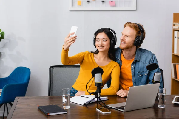 Asian broadcaster in wireless headphones taking selfie on smartphone with young colleague — Stock Photo