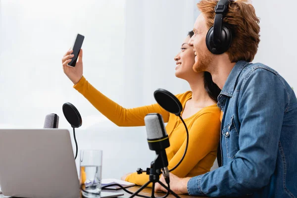 Selective focus of asian broadcaster taking selfie on smartphone with excited colleague in radio studio — Stock Photo