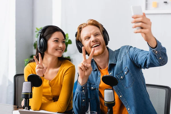 Excited, redhead announcer taking selfie on mobile phone while showing victory signs together with asian colleague — Stock Photo