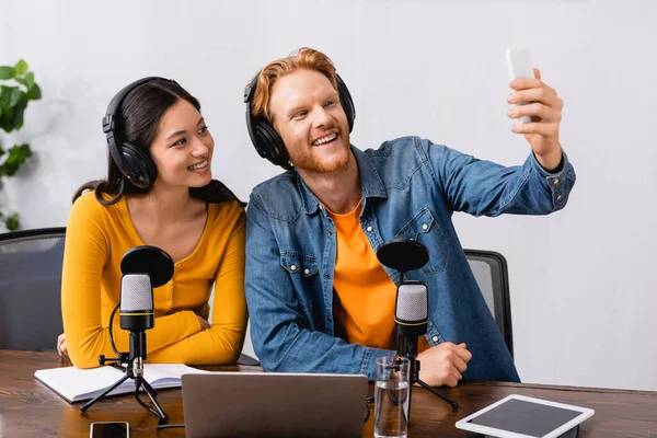 Joven freelancer tomando selfie de smartphone junto con asiático colega en auriculares inalámbricos - foto de stock