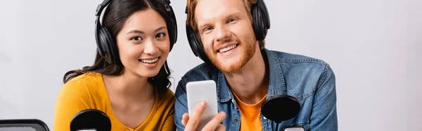 Concept panoramique de rousse hôte radio tenant smartphone tout en regardant la caméra avec un collègue asiatique — Photo de stock