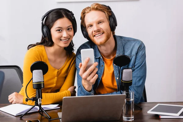 Jeune radiodiffuseur tenant smartphone tout en regardant la caméra avec un collègue asiatique en studio de radio — Photo de stock