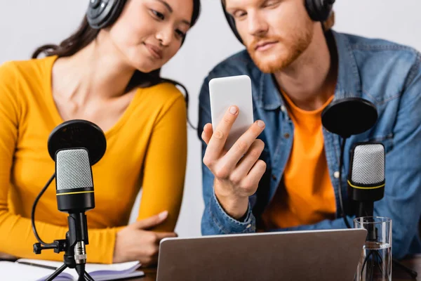 Selective focus of young broadcaster using smartphone near asian colleague in wireless headphones — Stock Photo