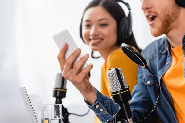 Aufgeregter Ansager mit Smartphone nahe asiatischem Kollegen in drahtlosem Kopfhörer — Stockfoto