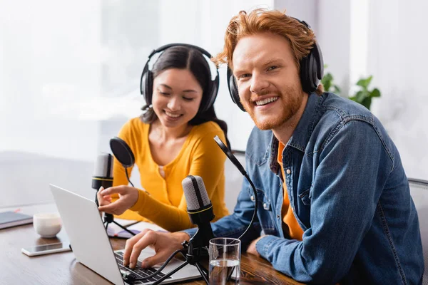 Selective focus of asian broadcaster pointing with finger at laptop near excited colleague in wireless headphones — Stock Photo