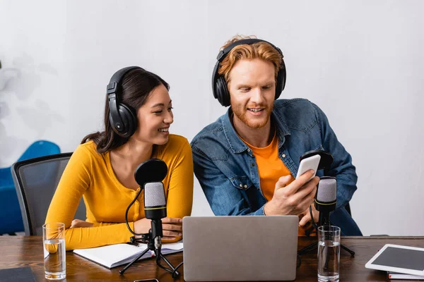 Joven locutor en auriculares inalámbricos utilizando teléfono inteligente cerca de asiático colega - foto de stock