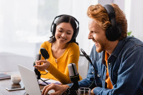 Selective focus of asian radio host pointing with finger at laptop near excited colleague in studio — Stock Photo