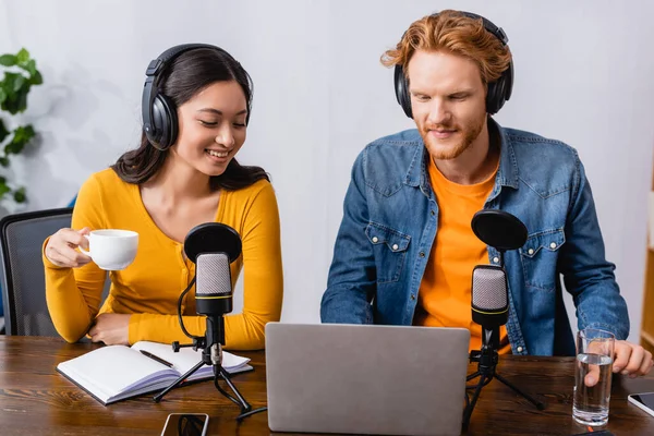 Asian broadcaster in wireless headphones holding cup of coffee near colleague in radio studio — Stock Photo
