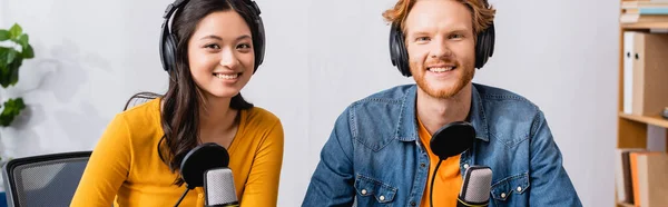 Panoramic concept of excited interracial couple of broadcasters in wireless headphones looking at camera in radio studio — Stock Photo