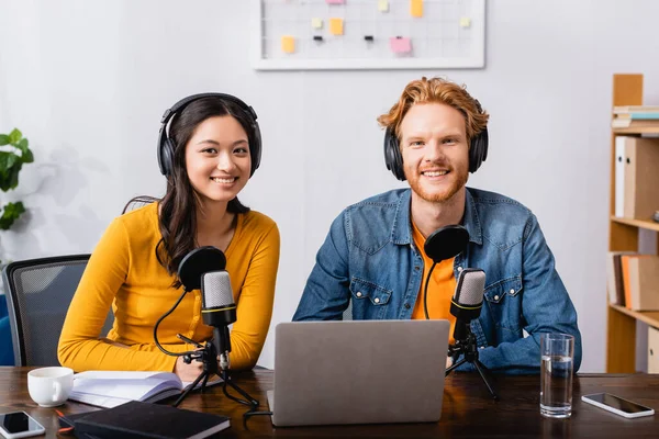 Young radio hosts in wireless headphones looking at camera near microphones in studio — Stock Photo