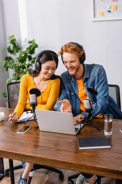 Giovane emittente che punta con mano al computer portatile vicino collega asiatico in cuffie wireless — Foto stock