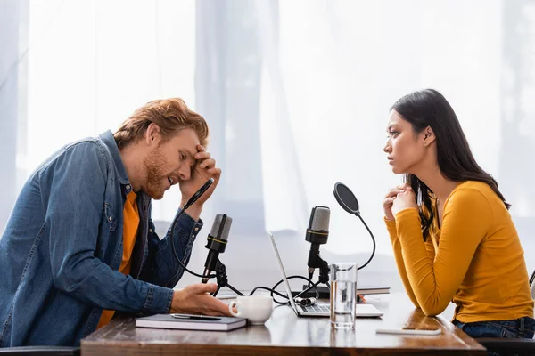 Joven asiático radio host mirando tenso hombre tocando cabeza durante entrevista en estudio - foto de stock