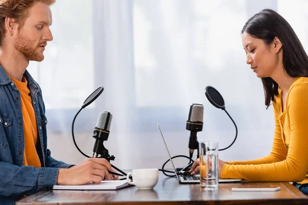 Side view of asian broadcaster using laptop, and redhead man looking at notebook during interview in studio — Stock Photo