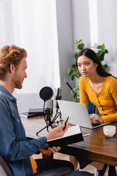 Redhead man holding notebook and pen while talking to young asian interviewer in studio — Stock Photo