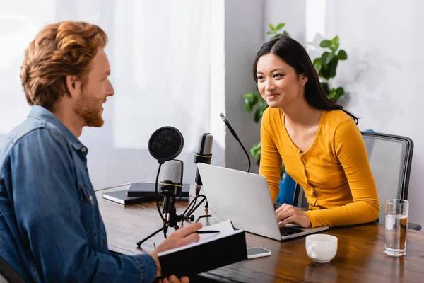 Young asian broadcaster using laptop while interviewing redhead man holding notebook and pen in studio — Stock Photo
