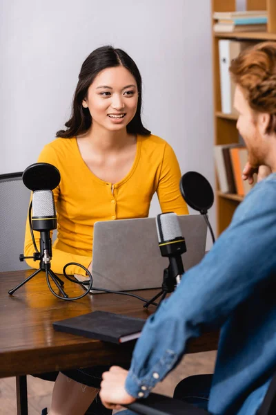 Selektiver Fokus eines jungen asiatischen Radiomoderators mit Laptop während eines Interviews mit einem Mann im Studio — Stockfoto
