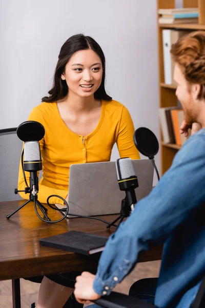 Foco seletivo de jovem asiático rádio host usando laptop enquanto entrevistando homem em estúdio — Fotografia de Stock