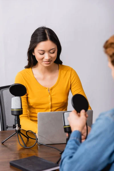 Selective focus of young asian radio host using laptop during interview with man in studio — Stock Photo