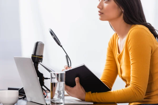 Selective focus of asian broadcaster holding notebook and pen near microphone at workplace — Stock Photo