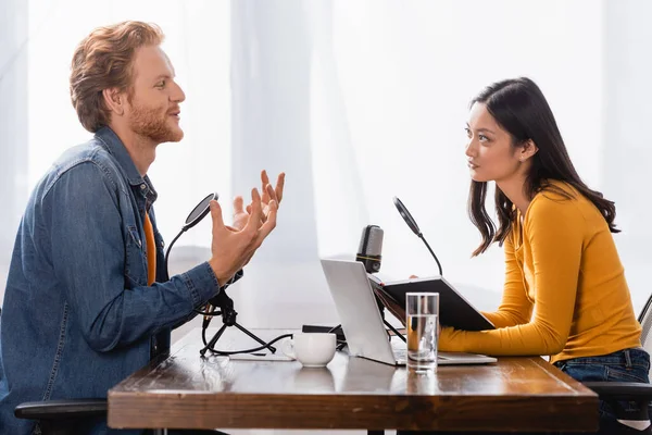 Side view of redhead man gesturing while talking to young asian radio host in studio — Stock Photo