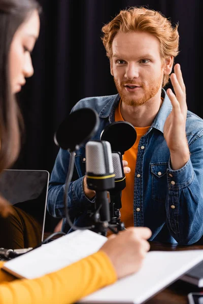 Вибірковий фокус рудого чоловіка жестикулює під час інтерв'ю з молодим азіатським радіоведучим, що пише в блокноті — стокове фото