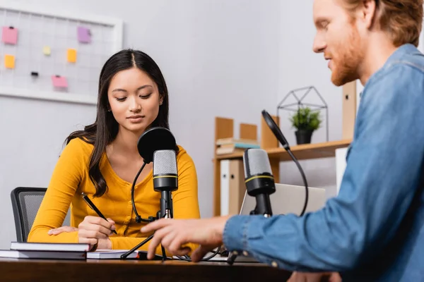Selective focus of young asian broadcaster holding pen during interview with redhead man — Stock Photo