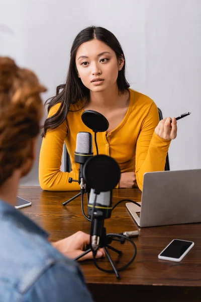Foco seletivo de morena asiática emissora segurando caneta durante entrevista com o homem no estúdio de rádio — Fotografia de Stock