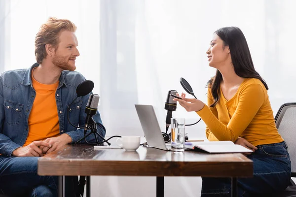 Brunette asian broadcaster holding pen while talking to young man in radio studio — Stock Photo