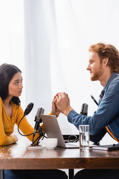 Young redhead radio host holding hands with sad asian woman during interview in studio — Stock Photo