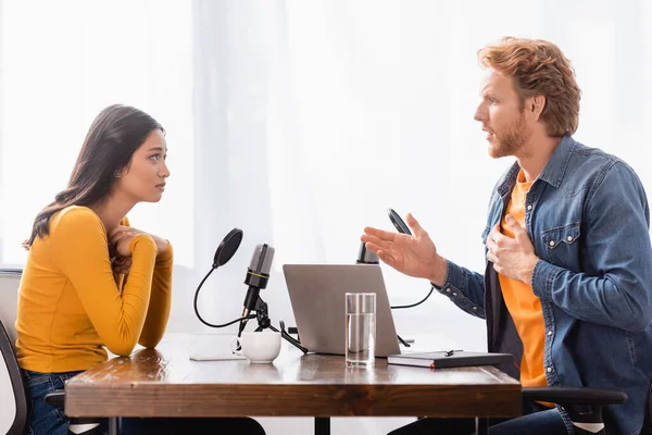 Side view of interviewer gesturing while talking to shocked asian woman holding hands near chest — Stock Photo