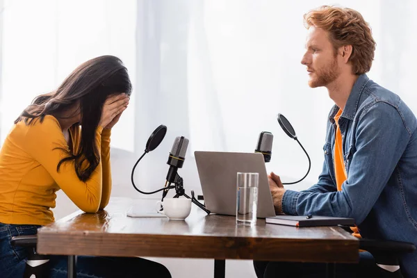 Side view of redhead broadcaster looking at stressed brunette woman obscuring face with hands during interview — Stock Photo