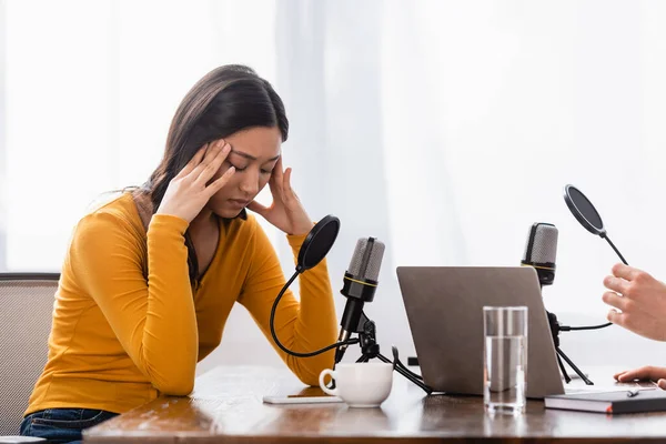 Interviewer near upset asian woman with closed eyes touching head in radio studio — Stock Photo